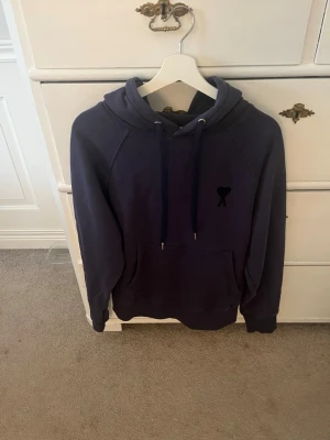 Mörkblå hoodie med hjärtdetalj - Snygg mörkblå hoodie med huva och dragsko samt en svart hjärtdetalj på bröstet. Hoodien har en klassisk känguruficka framtill och långa ärmar. Perfekt för en chill och avslappnad stil.