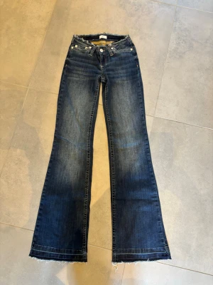 Mörkblå bootcut jeans från Nelly - Säljer ett par mörkblå bootcut jeans med uppsträttade kanter nertill. Jeansen har klassisk femficksmodell, låg midja och snygg tvätt med ljusare partier framtill och baktill. Perfekta för en avslappnad och trendig look. Använda en gång innan.