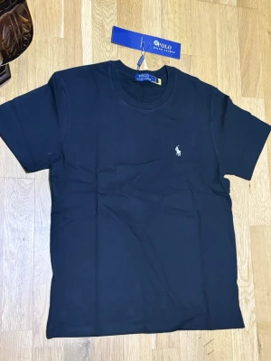 Svart t-shirt från Polo Ralph Lauren - Klassisk svart t-shirt från Polo Ralph Lauren med rund halsringning och korta ärmar. Liten vit broderad logga på bröstet ger en clean och stilren look. Perfekt basplagg till garderoben.