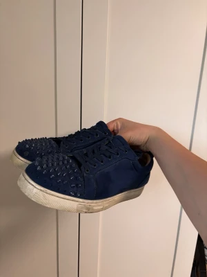 Blå sneakers med nitar och röd sula - Unika blå sneakers med coola nitar över hela tån och sidan. Skorna har klassisk röd sula och är tillverkade i mocka. Perfekta för dig som vill sticka ut och addera lite edge till din outfit.