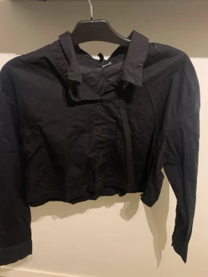 Svart croppad skjorta med krage - Säljer en svart croppad skjorta med klassisk krage och knappar framtill. Skjortan har långa ärmar och en loose passform som ger en avslappnad vibe. Perfekt att styla med höga jeans eller kjol.