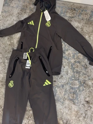 Grå Real Madrid träningsset Adidas - Snyggt grått träningsset från Adidas x Real Madrid med neongula detaljer. Setet består av en hoodie med dragkedja och två fickor samt matchande byxor med resår i midjan och dragkedjor vid fickorna. Perfekt för fotbollsträning eller chill. Helt ny perfekt inför sommaren 