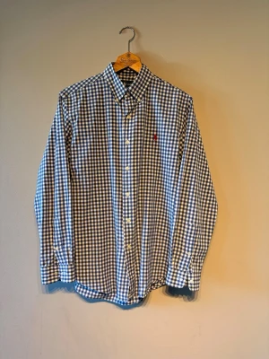 Blårutig skjorta från Polo Ralph Lauren - Säljer en klassisk blårutig skjorta från Polo Ralph Lauren med den ikoniska röda broderade loggan på bröstet. Skjortan har lång ärm, knappar framtill och är tillverkad i mjuk bomull. Perfekt för dig som gillar tidlös stil.