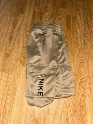 Guldfärgade cargobyxor från Nike - Snygga guldfärgade cargobyxor från Nike med elastisk midja och muddar vid benslut. Byxorna har stora cargofickor på sidorna och en diskret Nike-logga på vänster ben. Tillverkade i ett mjukt och glansigt material som ger en sportig look. Storlek S