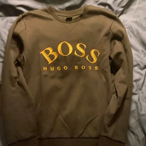 Svart sweatshirt från Hugo Boss - Säljer en snygg svart sweatshirt från Hugo Boss med stort gult BOSS-tryck på framsidan. Tröjan har långa ärmar och en klassisk rund halsringning. Perfekt för en stilren och bekväm look.
