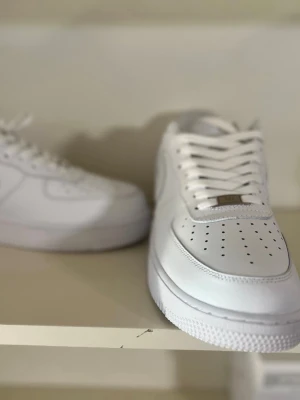 Vita Nike Air Force 1 sneakers - Klassiska helvita Nike Air Force 1 sneakers i läder med perforerad tåbox, vita skosnören och metallbricka på snörningen. Swoosh-logga på sidan och tjock sula för en clean och tidlös look.