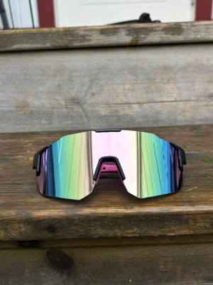 Pastellrosa / svart ram Sportiga spegelglasade solglasögon - Säljer ett par sportiga solglasögon med spegelglas i regnbågsfärger och svart båge. Glasen har en futuristisk, kantig form och ger en cool vibe till din outfit. Perfekta för dig som vill sticka ut och gillar en modern look.