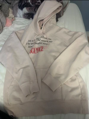 Rosa hoodie med Scarface-tryck - Säljer en rosa hoodie med huva och känguruficka. På bröstet står det 'He was Tony Montana. The world will remember him by another name SCARFACE' i svart och rött tryck. Hoodien har ribbade muddar och dragsko i huvan. Perfekt för dig som gillar filmreferenser.