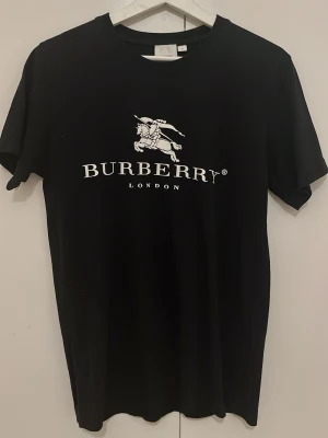 Svart Burberry t-shirt med tryck - Svart t-shirt från Burberry med klassiskt vitt logotryck och riddarmotiv på bröstet. T-shirten har rund hals och korta ärmar, perfekt för en stilren och trendig look.