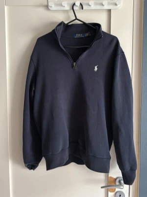 Mörkblå halvzip från Polo Ralph Lauren - Mörkblå långärmad tröja från Polo Ralph Lauren med halv dragkedja och klassisk broderad logga på bröstet. Tröjan har en avslappnad passform och ribbade muddar vid ärmslut och nederkant. Perfekt för en stilren och bekväm look.