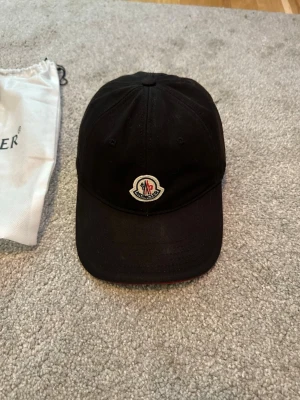 Svart keps från Moncler - Tja säljer en Stilren svart keps från Moncler med klassisk böjd skärm och broderad logotyp framtill. Justerbar rem baktill med röd, vit och blå detalj samt metallspänne med Moncler-gravyr. Perfekt nu till sommaren 