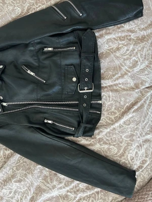 Svart biker skinnjacka med bälte - Cool svart skinnjacka i klassisk bikerstil med flera silvriga dragkedjor, axelklaffar och bälte i midjan. Jackan har snedställda fickor och många detaljer. Perfekt för dig som vill ha tuff stil.
