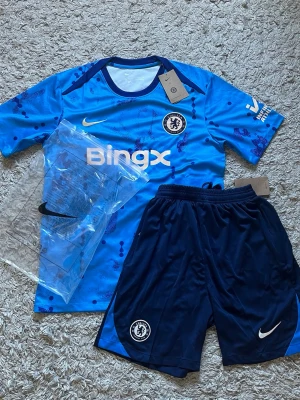 Chelsea FC Nike fotbollskit blå [FAST PRIS] - Chelsea FC matchställ från Nike med blå tröja och mörkblå shorts. Tröjan har Bingx-logga, klubbmärke och Nike Swoosh, samt mönster i olika blå nyanser. Shortsen har elastisk midja och klubbmärke. Perfekt för fotbollsträning eller match.