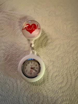 Vit sjuksköterskeklocka med hjärtmotiv - Vit sjuksköterskeklocka från Tempo med clipsfäste och ett hjärtmotiv med EKG-linje på ovandelen. Klockan har tydliga siffror och röda visare, perfekt att fästa på arbetskläder. Praktisk och stilren accessoar för dig som jobbar inom vården.