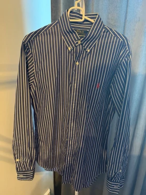 Randig skjorta från Polo Ralph Lauren - Snygg blå skjorta med vita ränder från Polo Ralph Lauren. Slim fit-modell med klassisk krage och knappar framtill. Ikonisk röd logga broderad på bröstet. Perfekt för en stilren look.