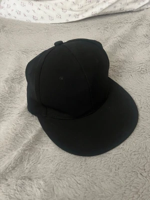 Svart snapback keps - Snygg svart snapback keps med rak skärm och klassisk design. Kepsen är helt svart utan logga eller tryck och har justerbar baksida. Tillverkad i bomullsmix för en bekväm passform. Perfekt för en clean och stilren look.