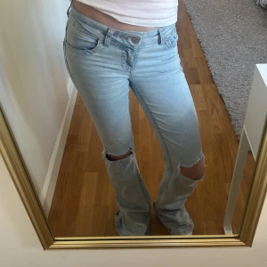 Ljusblå bootcut jeans med hål - Säljer ett par ljusblå bootcut jeans från Nelly med slitna detaljer vid knäna och broderade bakfickor. Jeansen har klassisk femficksdesign och låg midja. Perfekta för en avslappnad och trendig look.