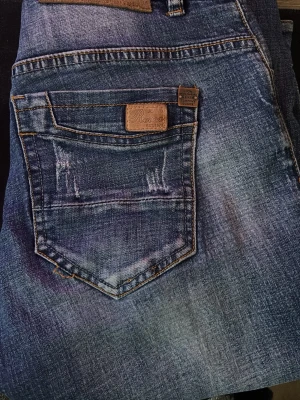 Blå slitna jeans från Revox - Säljer ett par blå jeans från Revox med slitningar och distressed detaljer på både fram- och baksida. Jeansen har klassisk femficksdesign, raka ben och tydliga tvättade partier för en cool look. Materialet är denim i bomull.