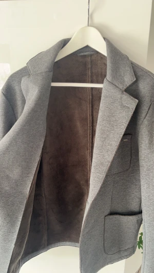 Stickad kavaj med foder - Stilren grå kavaj med mjukt brunt foder på insidan. Perfekt för en avslappnad men ändå uppklädd look. Den är fodrad på hela insidan, med andra ord är den otroligt skön. Passar M/L