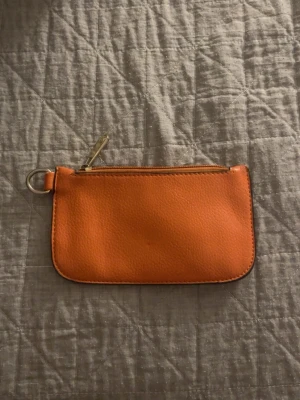 Orange plånbok Michael Kors - Snygg orange plånbok i läderimitation med dragkedja och metallring i ena hörnet. Perfekt för kort, mynt och småsaker. Enkel och stilren design med rundade hörn och slät yta.