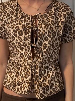 Leopardmönstrad blus med snörning - Snygg leopardmönstrad blus med korta ärmar och flera snörningar framtill. Modellen har en lätt utställd passform och markerad midja. Perfekt för dig som vill sticka ut med ett djurmönster.