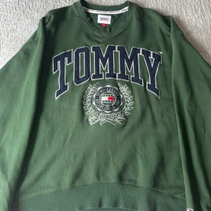 Grön sweatshirt från Tommy Hilfiger - Snygg grön sweatshirt från Tommy Hilfiger med stort TOMMY-tryck i marinblått och vitt framtill samt broderad logga och emblem. Klassisk rund halsringning och ribbade muddar vid ärmslut och nederkant. Perfekt för en avslappnad och trendig look.