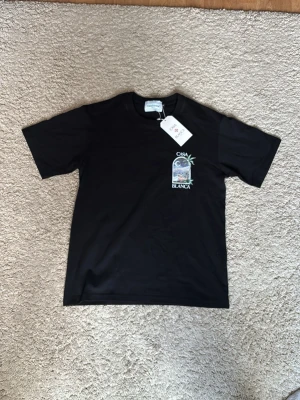 Svart t-shirt från Casablanca - Svart t-shirt från Casablanca med tryck framtill som visar ett fönster med palmer och stadsmotiv. Klassisk rund halsringning och korta ärmar. Perfekt för dig som gillar stilrena plagg med unik detalj.