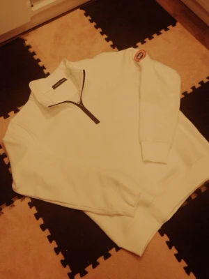 Vit halvzip tröja med patch - Vit stickad tröja från Weekday med halvzip och svart dragkedja. Tröjan har ribbade muddar och en rund patch på ena ärmen. Perfekt för lager på lager och enkel att styla till olika outfits.