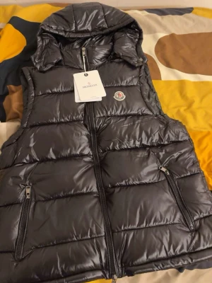 Svart dunväst från Moncler med huva - Snygg svart dunväst från Moncler med glansig finish och avtagbar huva. Västen har två dragkedjefickor framtill och klassisk Moncler-logga på bröstet. Perfekt för lager-på-lager och streetstyle.