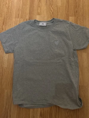 Grå t-shirt från Ami Paris - Säljer en stilren grå t-shirt från Ami Paris med det ikoniska hjärta-A broderiet på bröstet. T-shirten har rund halsringning och korta ärmar. Tillverkad i mjuk bomull för maximal komfort och enkel att matcha till olika outfits. Kom gärna med bud