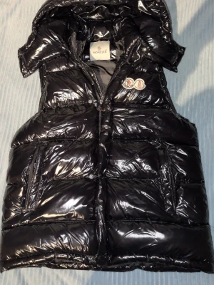 Svart glansig dunväst från Moncler - Snygg svart dunväst från Moncler med glansigt material och avtagbar huva. Västen har en klassisk Moncler-logga på bröstet och dragkedja framtill. Perfekt till sommaren och varmare väder. Den är lite sönder innuti skriv för bild men det syns inte och märks inte när man har på den på något sätt. Säljer pågrud av att jag inte andvänder den längre. Skriv för frågor och mer bilder. Storlek S men passar M.