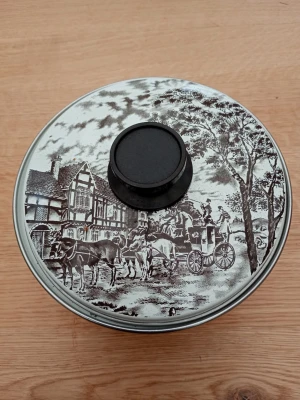 Kastrull med lock och vintage-motiv - Säljer en rund kastrull med lock, dekorerad med ett svartvitt vintage-motiv av häst och vagn framför ett gammalt hus och träd. Kastrullen har ett svart handtag på locket och passar perfekt för förvaring av småsaker eller kakor. Handtag fattas på sidan.