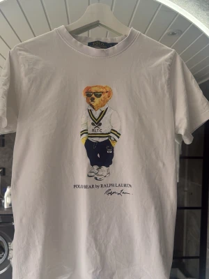Vit Polo Bear t-shirt från Ralph Lauren - Vit t-shirt från Ralph Lauren med ikoniskt Polo Bear-tryck framtill. T-shirten har rund hals, korta ärmar och är gjord i mjuk bomull. Perfekt för dig som gillar klassisk men lekfull stil. Skriv med de minsta fundering//William
