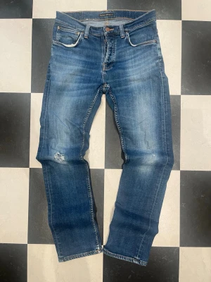 Nudie grim tim - Snygga blå jeans från Nudie Jeans med klassisk femficksdesign och orangea sömmar på bakfickorna. Jeansen har en lätt tvättad look och är tillverkade i denim. Perfekta för en avslappnad stil. Jeansen har slitningar på båda knän