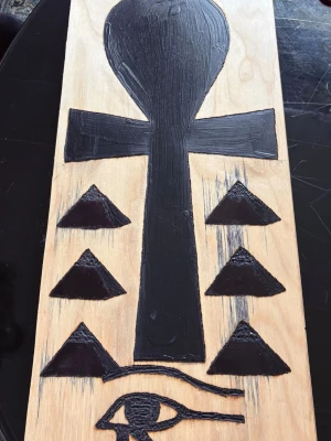 Träplatta med egyptiska symboler - Unik träplatta med handmålade egyptiska symboler, inklusive ett stort ankh-kors i guld och svart, pyramider och Horus öga. Plattan är rektangulär och har en naturlig träfärg som bas. Perfekt som dekoration för dig som gillar mystik och historia.
