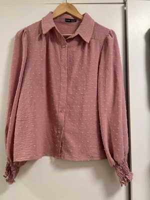Rosa blus  - Söt rosa blus med diskret strukturmönster. Blusen har klassisk krage, knäppning framtill och långa ärmar med smockade muddar. Perfekt för en trendig och feminin look.