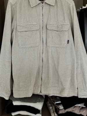 Beige overshirt i manchester - Stilren beige overshirt från ONLY & SONS i mjuk manchester. Jackan har två stora bröstfickor med lock, dragkedja framtill och klassisk krage. Perfekt för lager-på-lager och ger en avslappnad men trendig look.