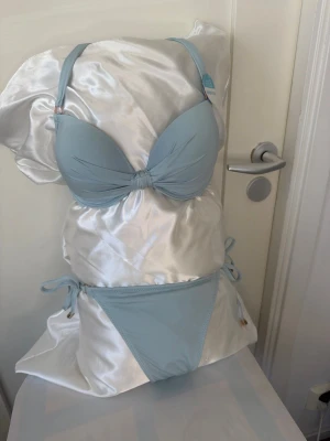 Ljusblå bikini med knyt och bygel - Snygg ljusblå bikini från Chantelle med vadderad bygel-bh och knyt i sidorna på bikinitrosan. Toppen har justerbara axelband och dekorativ knut framtill. Materialet är slätt och stretchigt, perfekt för stranden eller poolen.