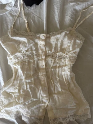 Ljusgult sommar linne med spets och knappar - Ljusgult sommar linne med smala axelband, dekorativa spetsdetaljer längs kanterna och bysten samt knäppning framtill. Tillverkat i ett tunt och luftigt material som känns som bomull eller linne. .