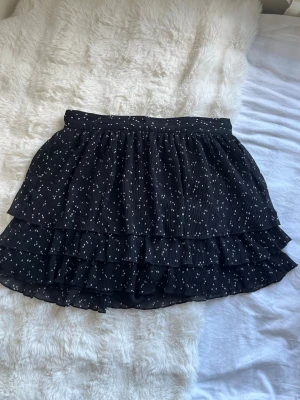 Svart volangkjol med små vita prickar - Snygg svart kortkjol med volanger och små vita prickar. Kjolen har flera lager och ett lätt, luftigt tyg som ger en skön känsla. Inbyggda shorts, liten i storlek.