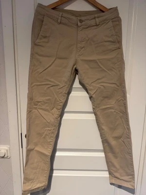 Beiga chinos med slim fit - Snygga beiga chinos med slim fit och klassisk design. Byxorna har snedställda fickor fram, två bakfickor och bälteshällor. Perfekta för en clean och stilren look. Tillverkade i mjukt bomullstyg för skön känsla hela dagen.