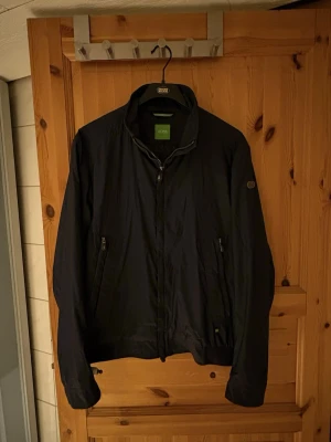 Svart vindjacka från Hugo Boss - Stilren svart vindjacka från Hugo Boss med dragkedja framtill och två fickor med dragkedja. Jackan har en klassisk krage och diskret logotyp på ärmen. Perfekt för blåsiga dagar och enkel att matcha med olika outfits.