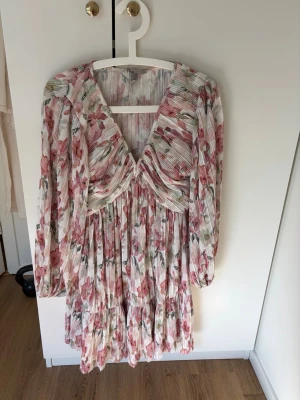 Blommig klänning med v-ringning - Superfin kortklänning med v-ringning och långa ärmar. Klänningen har ett plisserat tyg med blommigt mönster i rosa, grönt och vitt. 