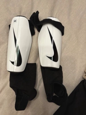 Nike benskydd vita med svart logga - Säljer ett par vita benskydd från Nike med svart Swoosh-logga. Skydden har elastiska svarta remmar och vadderad insida för extra komfort. Perfekta för fotbollsträning eller match. Lätta och smidiga att ta på.