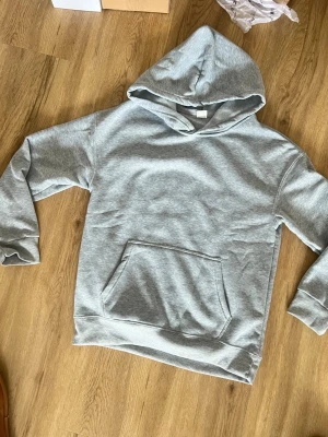 Grå hoodie med huva och ficka - Säljer en klassisk grå hoodie med stor huva och en praktisk magficka. Tröjan har ribbade muddar vid ärmslut och nederkant, perfekt för en avslappnad och cool look. Materialet är mjukt och bekvämt, idealiskt för chill dagar. Skick: ny , storlek: S 
