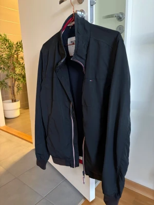 Svart vindjacka från Tommy Hilfiger - Snygg svart vindjacka från Hilfiger med diskret logga på bröstet och dragkedja med röd, vit och blå detalj. Jackan har hög krage, muddar vid ärmslut och meshfoder på insidan för extra komfort. Perfekt för blåsiga dagar och enkel att matcha.