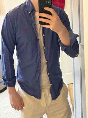 Blå skjorta från Ralph Lauren - Klassisk blå skjorta från Ralph Lauren med button-down krage och lång ärm. Skjortan har vita knappar och den ikoniska röda logotypen broderad på bröstet. Tillverkad i bomull för en bekväm och stilren look.