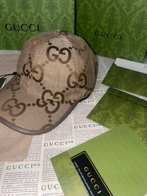 Beige keps med GG-mönster från Gucci - Snygg beige keps från Gucci med det klassiska GG-monogrammet i brunt över hela kepsen. Kepsen har böjd skärm och justerbar rem baktill. Tillverkad i canvasmaterial som ger en exklusiv känsla och streetig look.