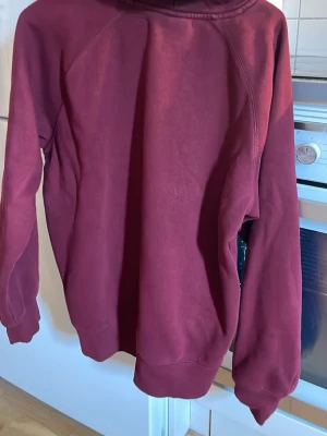 Vinröd hoodie från Carhartt - Säljer en vinröd hoodie från Carhartt med klassisk känguruficka och dragsko i huvan. Hoodien har ribbade muddar och en liten Carhartt-logga på ärmen. Perfekt för dig som gillar streetwear och vill ha en snygg och bekväm tröja.