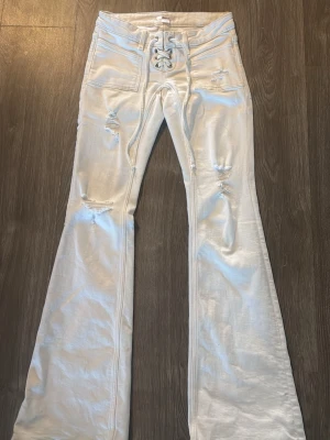 Vita utsvängda jeans med snörning - Andvända ungefär 3 gånger Säljer ett par vita jeans med utsvängda ben och trendiga slitningar. Jeansen har hög midja, snörning framtill och stora fickor både fram och bak. Perfekta för dig som vill sticka ut med en cool retrostil.